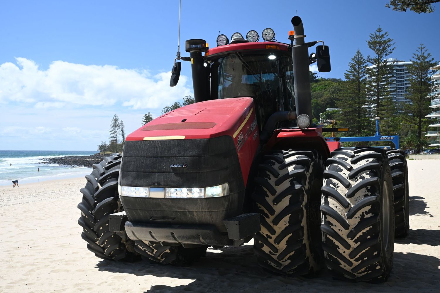 A large red Case IH tractor parked on a sandy beach with ocean waves and pine trees in the background.