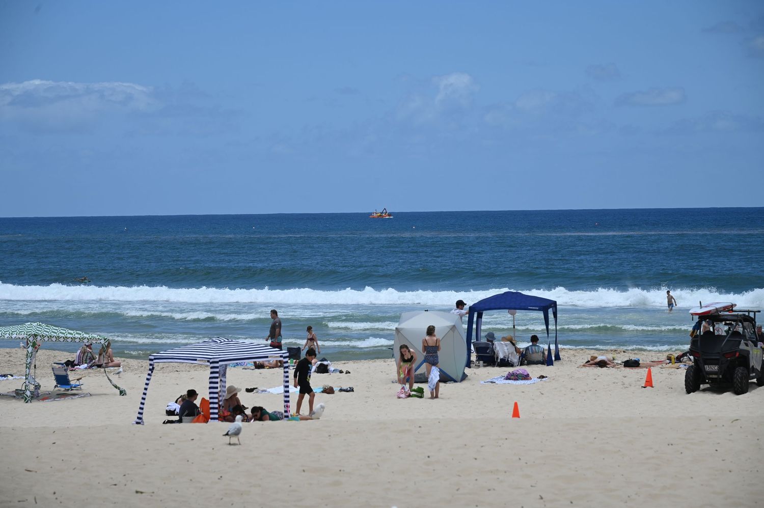 Beachgoers relax on a sunny sandy beach with blue ocean waves, a canopy tent, orange cones, and an ATV nearby.