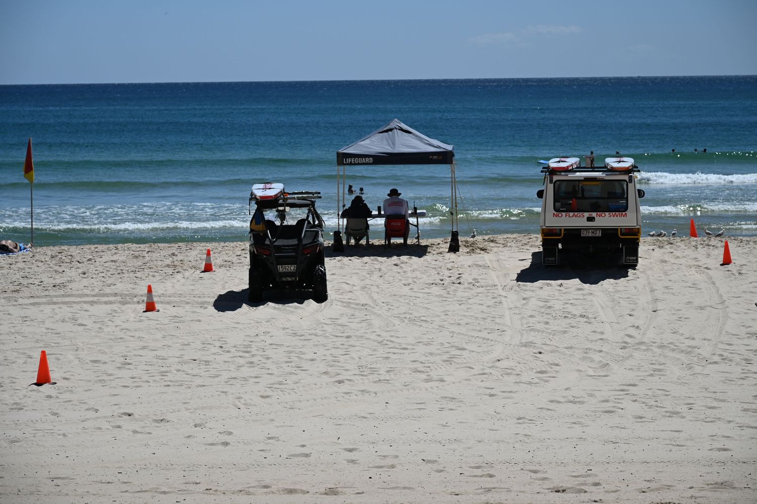 Beach patrol setup with orange cones, canopy tent, and vehicles on sandy shore near ocean waves.