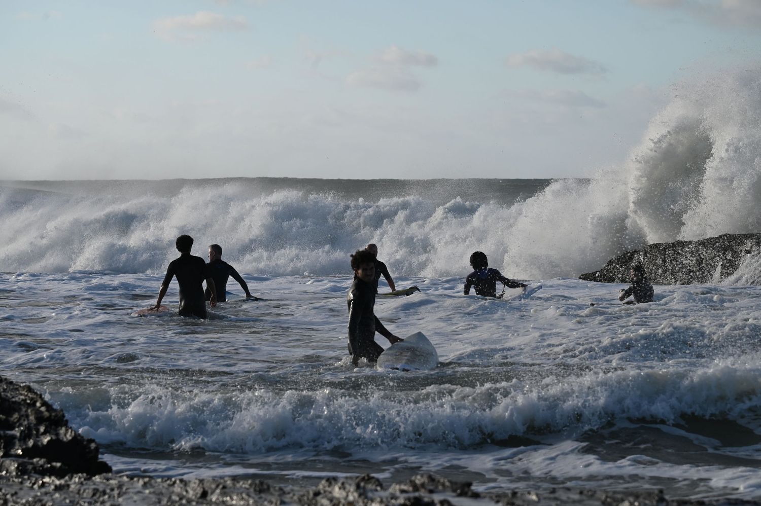 Silhouettes of people playing in rough ocean waves on a rocky shore during stormy weather.