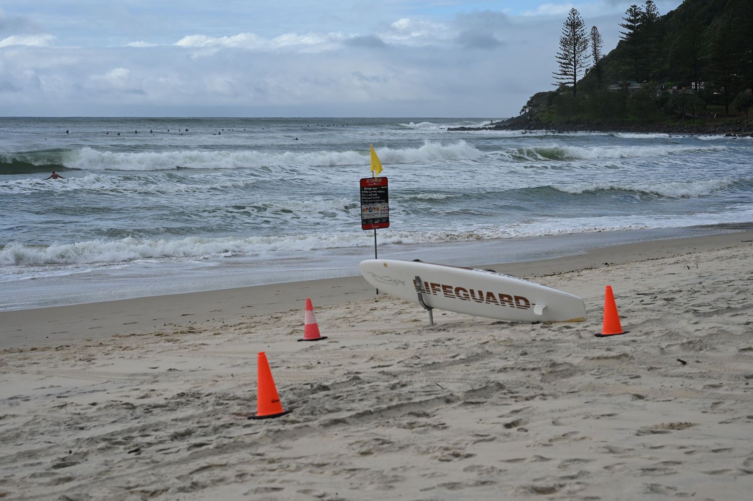 Lifeguard sign with orange cones on a sandy beach with rough waves and green headland in background.