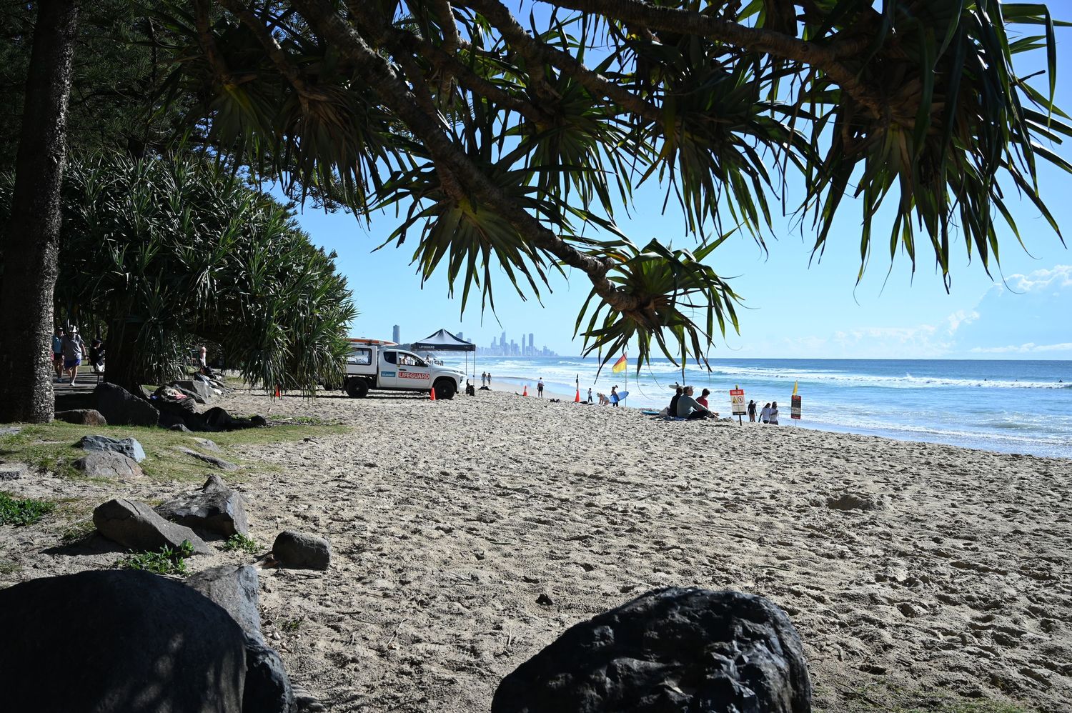 Sandy beach with palm trees, rocks, and people enjoying the ocean shoreline on a sunny day.