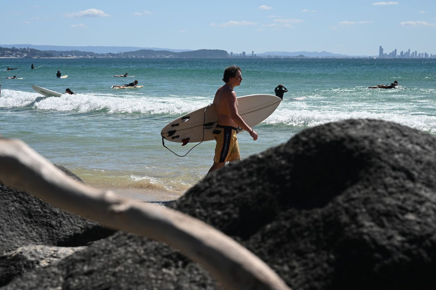 A surfer carrying a white surfboard walks along a rocky beach with ocean waves and a city skyline in the background.