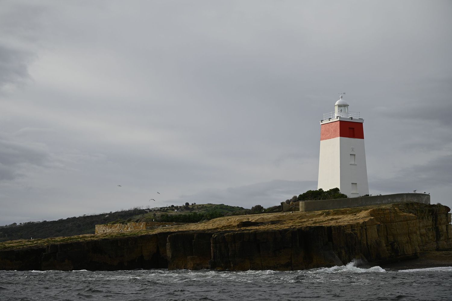 A white and red striped lighthouse stands on rocky coastal cliffs under a cloudy sky.