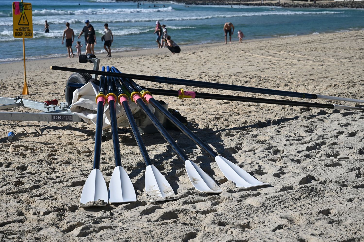 Rowing oars and blades stacked on sandy beach with people walking in background on sunny day.