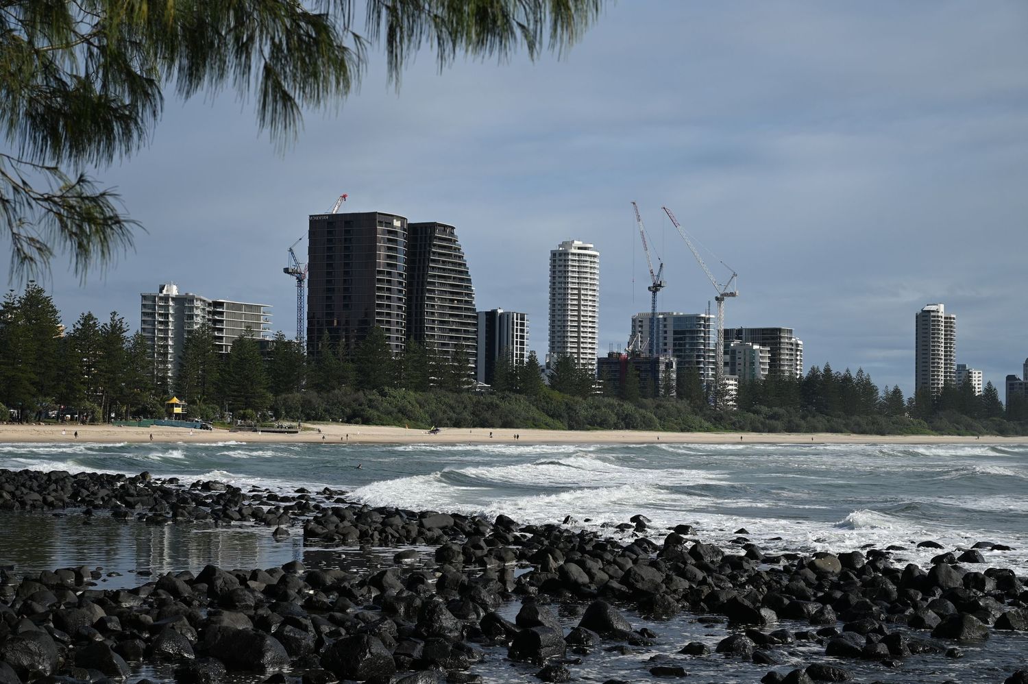 Rocky shoreline with crashing waves, lush trees, sandy beach, and city skyline with cranes under cloudy skies.