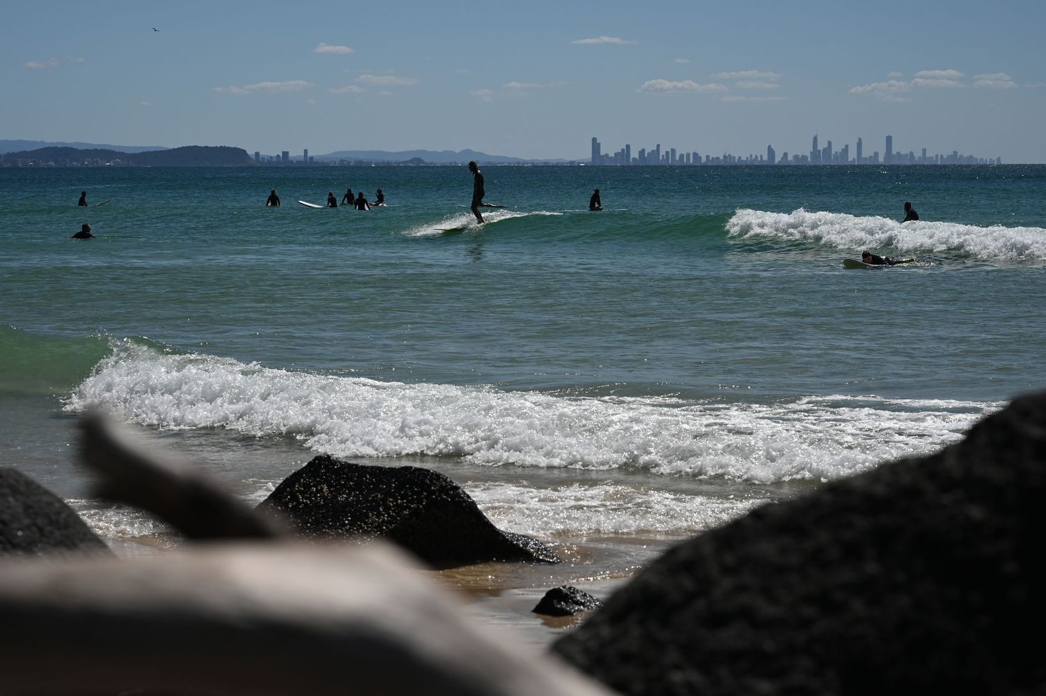 Surfers riding waves at a beach with a city skyline visible in the distance on a sunny day.