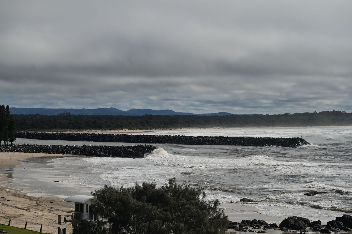 Stormy coastal beach scene with rocky breakwater, crashing waves, and overcast skies in Australia.