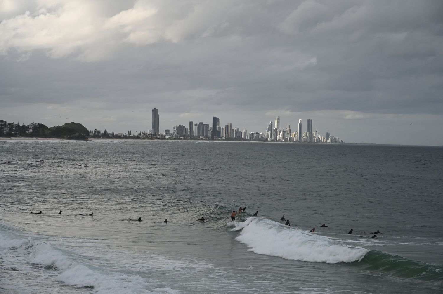 Surfers riding waves with dramatic Gold Coast city skyline under overcast skies in Australia.