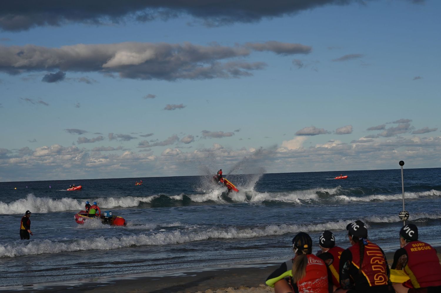 A surf rescue boat launching through breaking waves during a surf lifesaving competition on the beach.