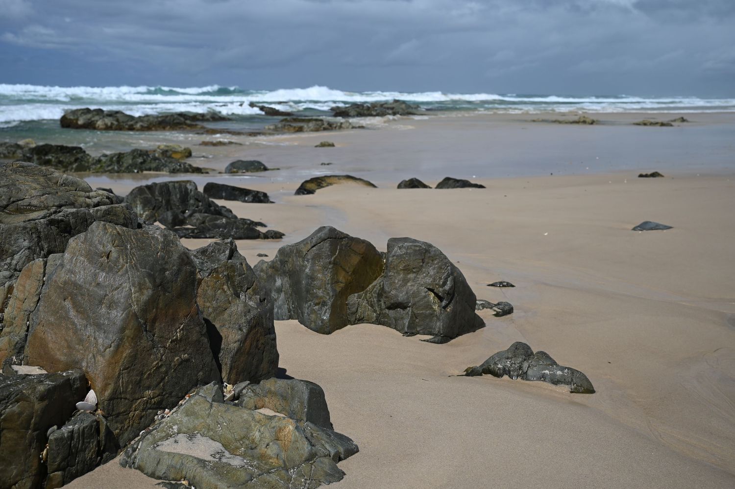 Rocky sandy beach with large boulders, golden sand, and stormy ocean waves under overcast skies.