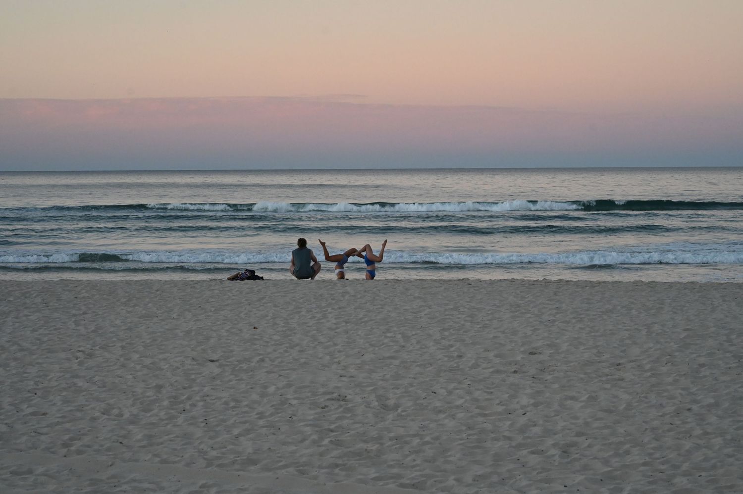 Person sitting with driftwood on a sandy beach at sunset with calm ocean waves.