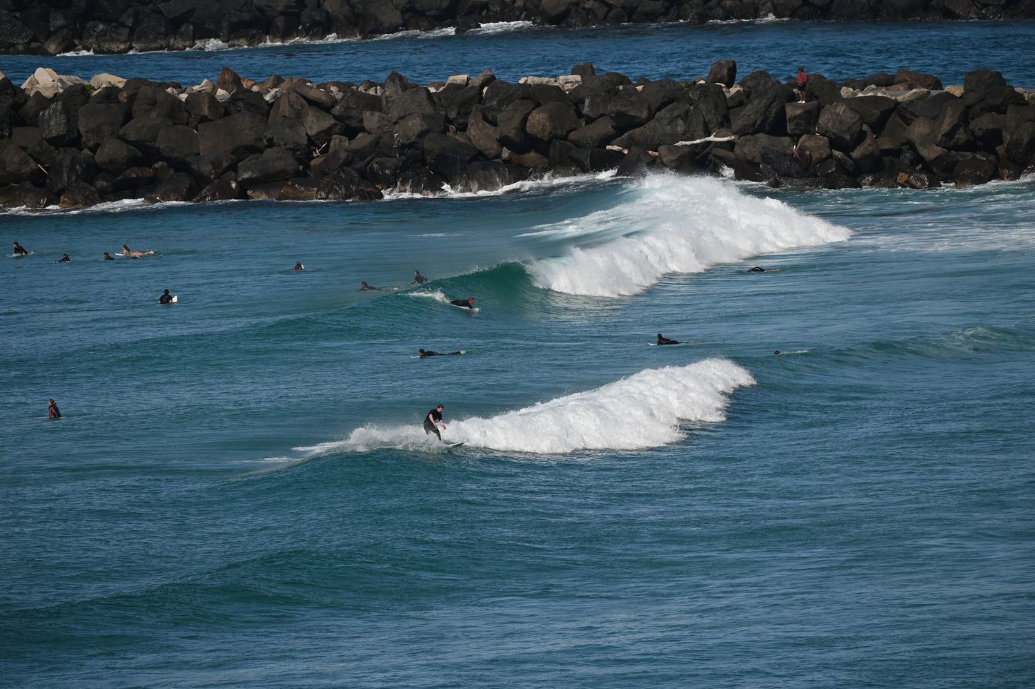 Surfers riding waves near a rocky breakwater in clear blue ocean waters on a sunny day.