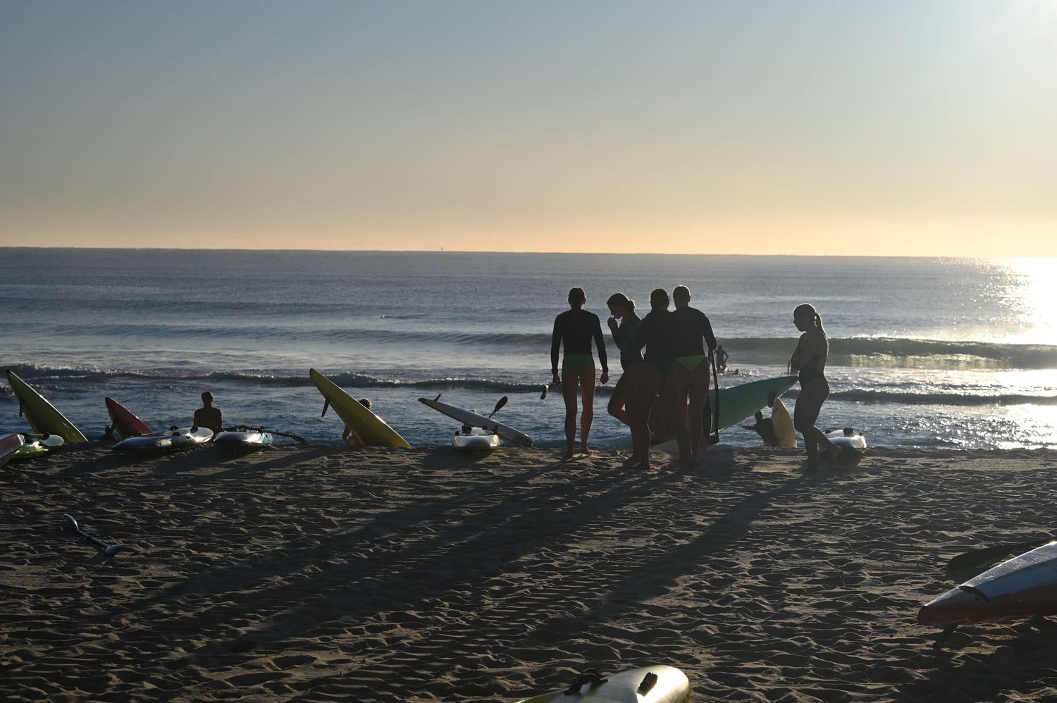 Silhouetted surfers gather on a sandy beach at sunset with surfboards, ocean waves in the background.