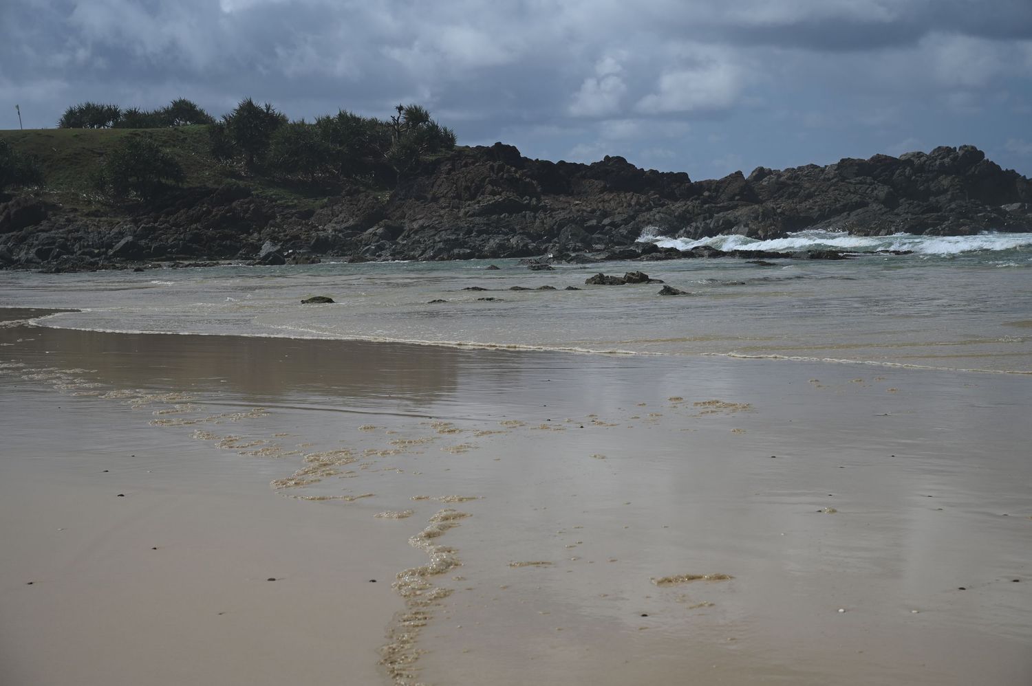 Sandy beach with rocky headland and crashing waves under cloudy skies on a overcast day.