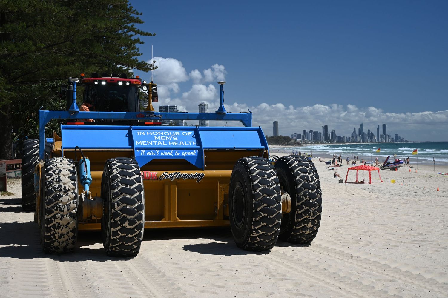 Large blue and yellow beach cleaning machine with oversized tires on sandy beach with city skyline in background.