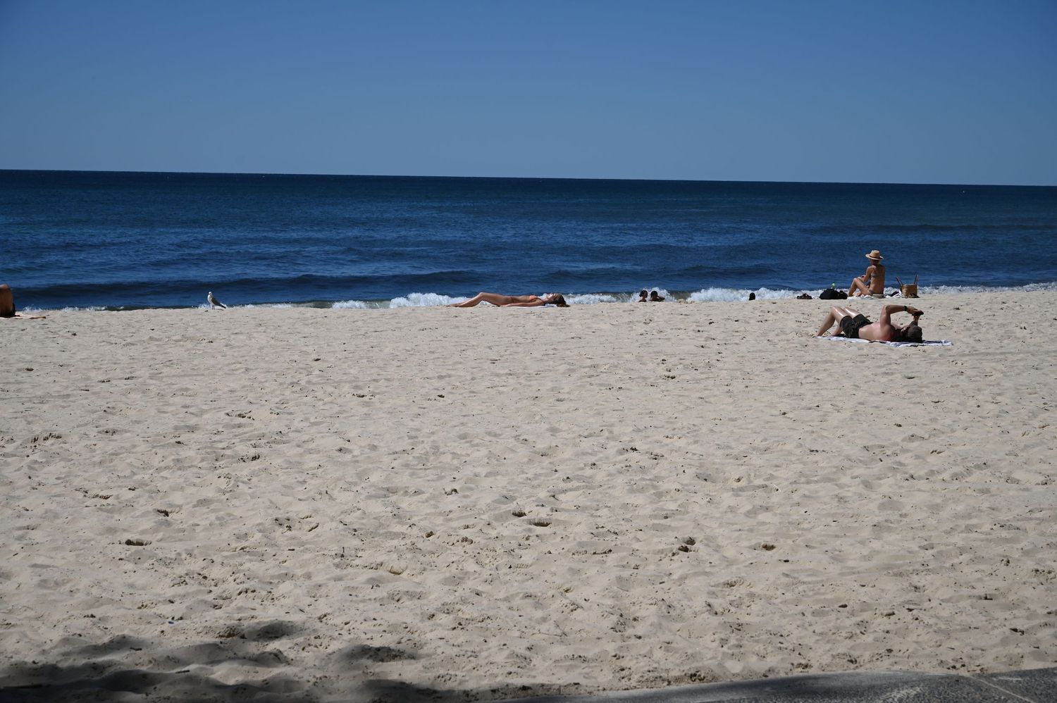 Sunny beach scene with white sand, calm ocean waves, and people relaxing on a clear blue sky day.