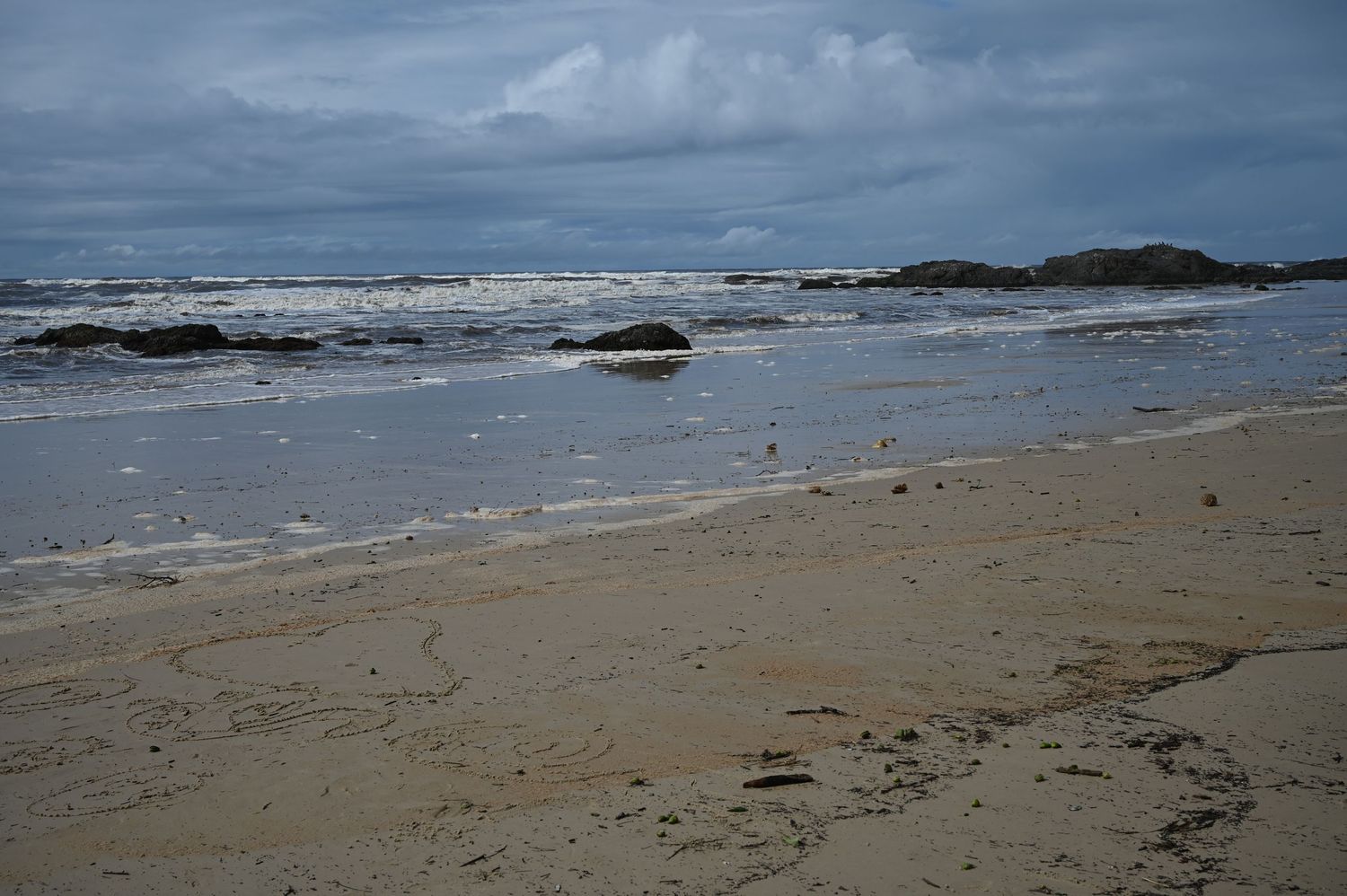 Cloudy coastal beach scene with rocky shoreline, gentle waves, and wet sand under overcast skies.