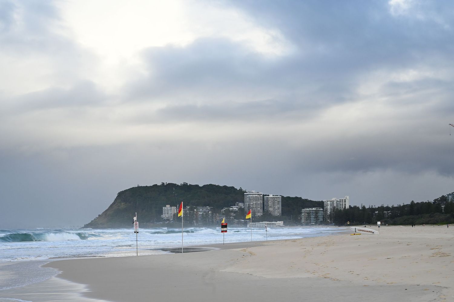 A wide sandy beach with surf lifesaving flags and a headland in the background under cloudy skies.
