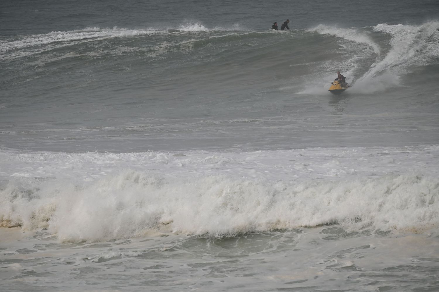 Surfer riding a large ocean wave on a gray stormy day with white foam crashing in the foreground.
