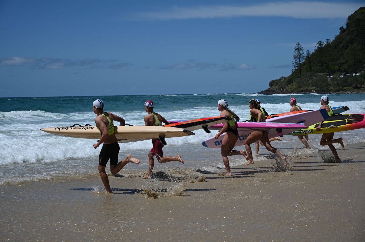 Surfers running into ocean waves carrying colorful surfboards during a beach competition on a sunny day.