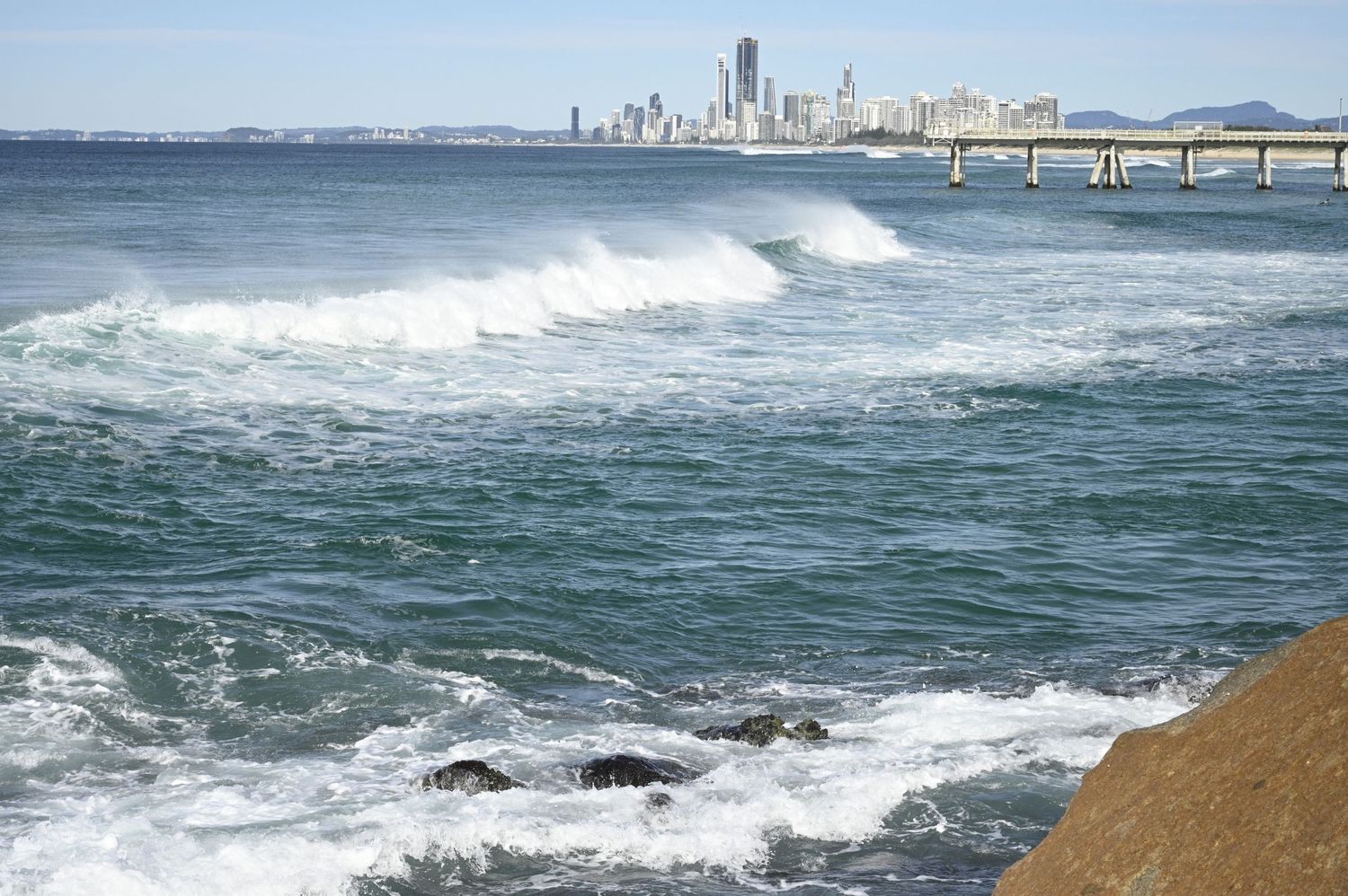 Waves crashing on rocky shore with city skyline and bridge in background on a clear day.