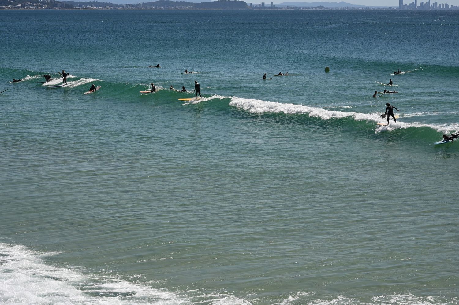 Surfers riding waves at a busy beach break with clear turquoise water on a sunny day.