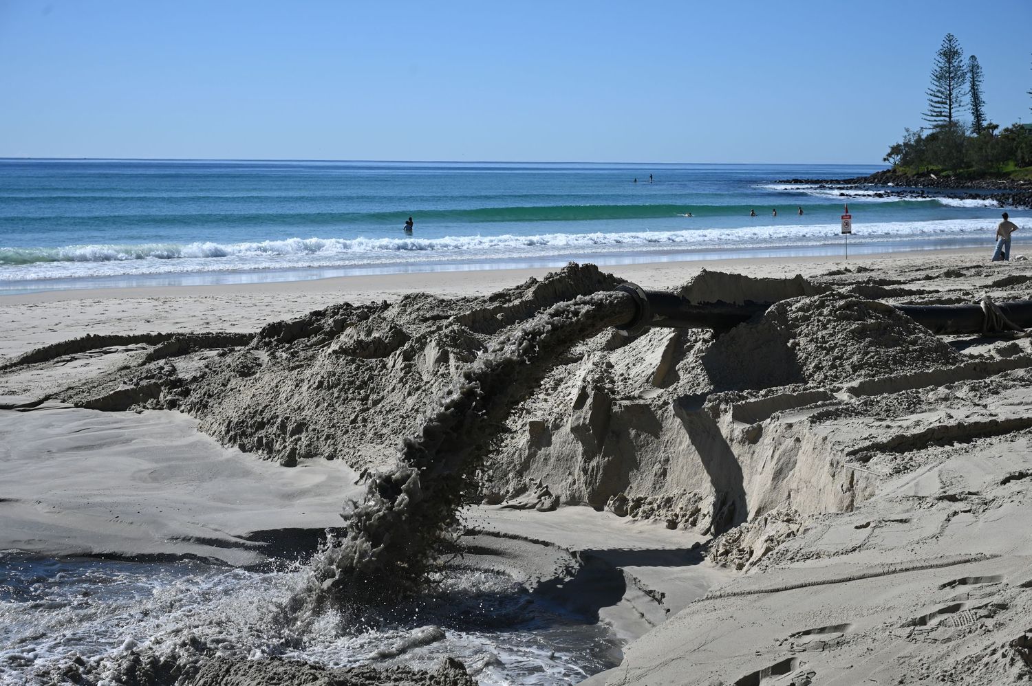 Eroded sand formations on a sunny Australian beach with turquoise ocean waves and pine trees in the background.