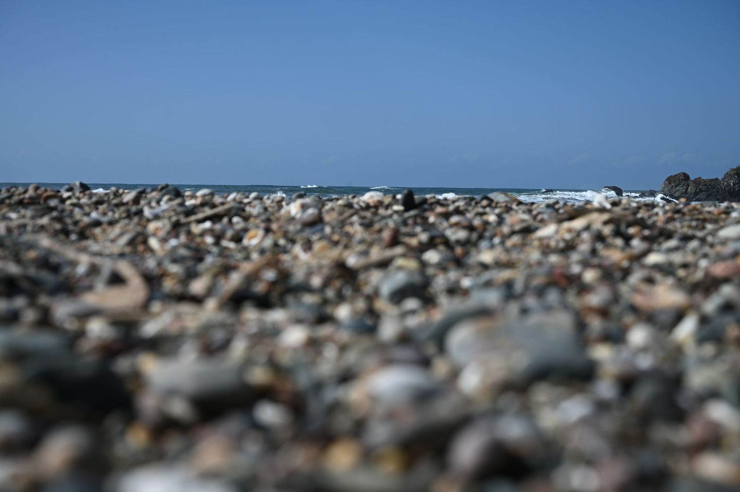 A rocky pebble beach stretches to the horizon under a clear blue sky on a sunny day.