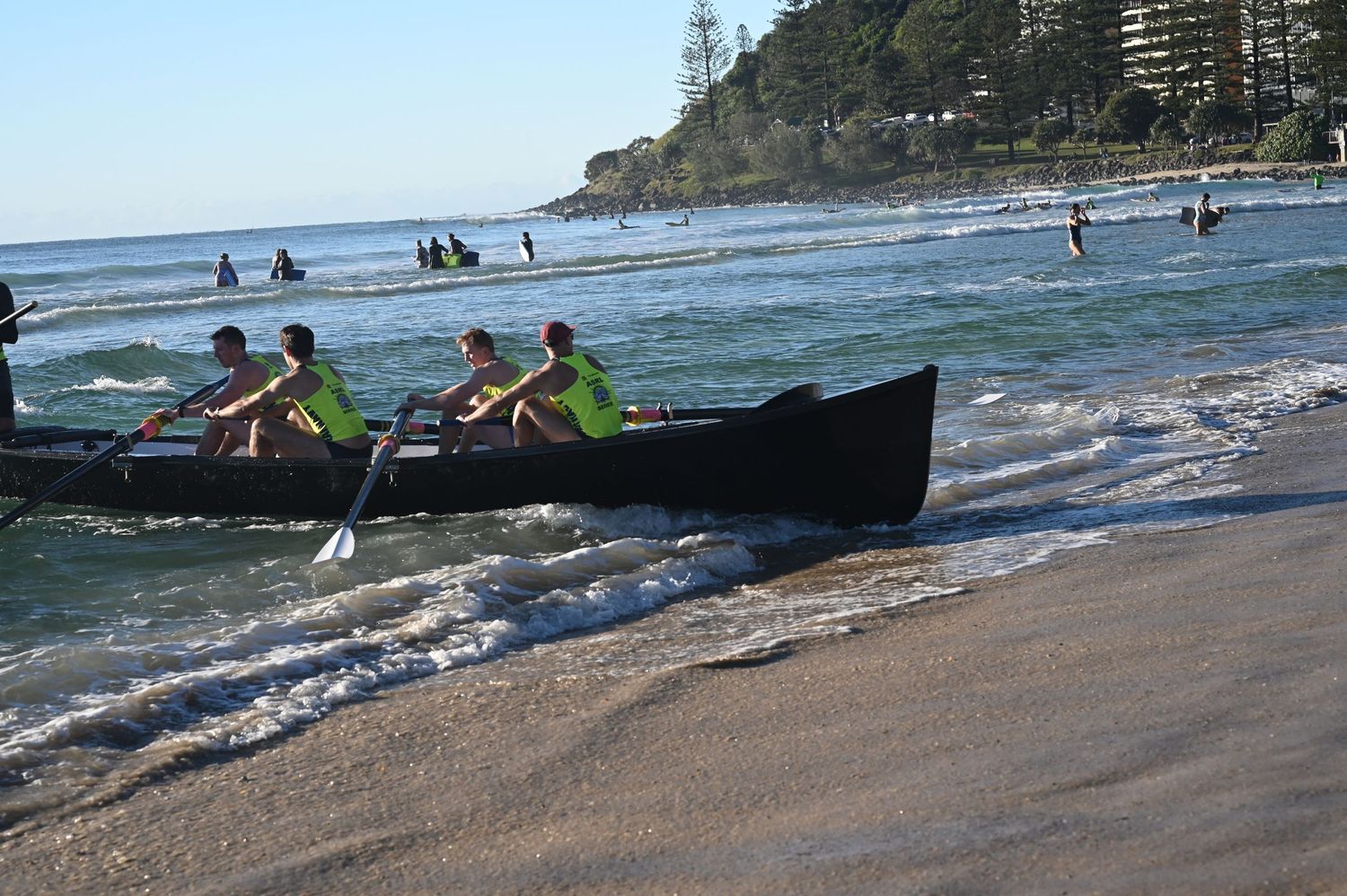 Team of rowers in green shirts paddle a long wooden canoe through shallow beach surf.