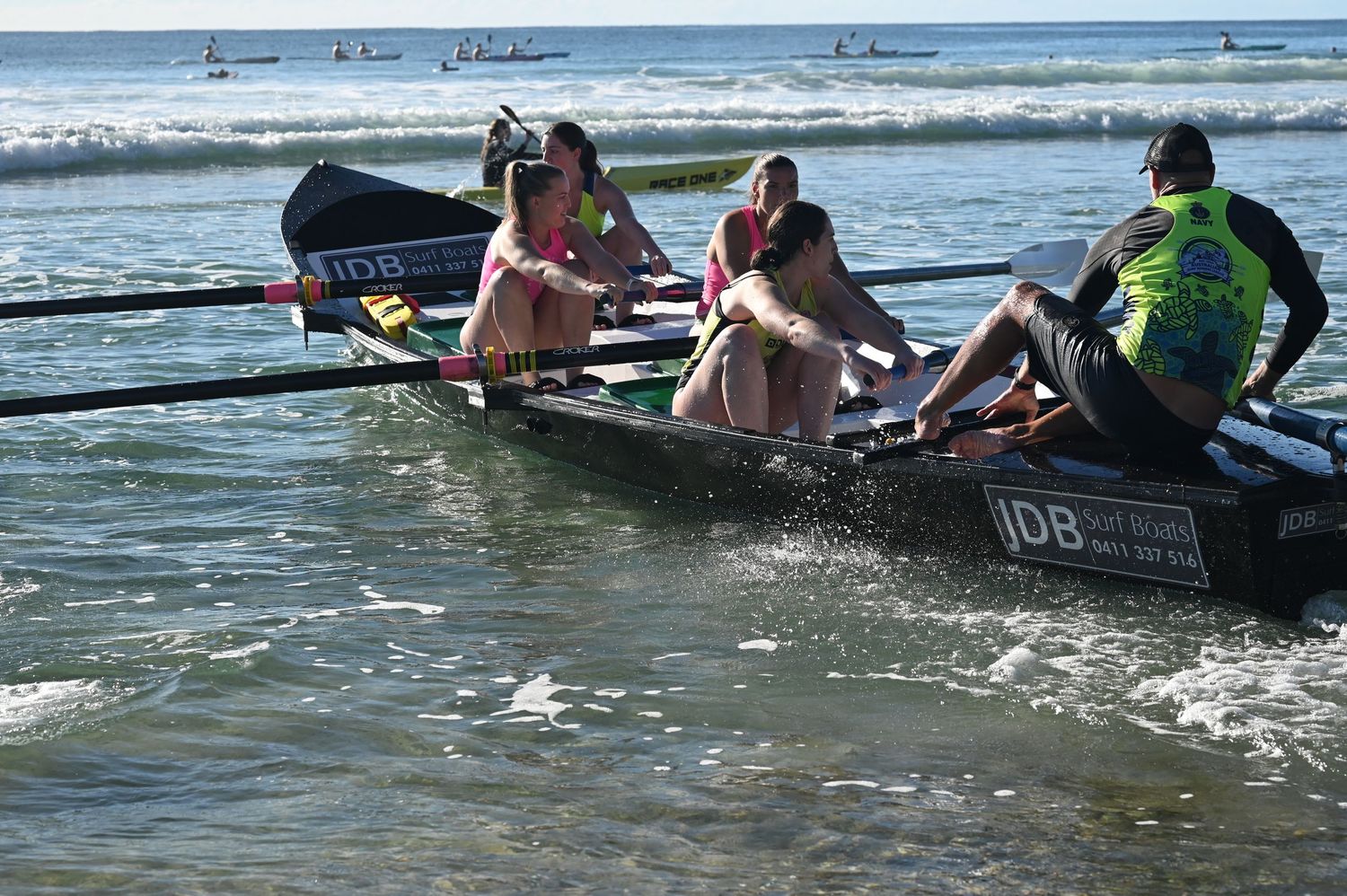 Surf lifesaving crew rows an IDB racing boat through ocean waves during a beach competition.