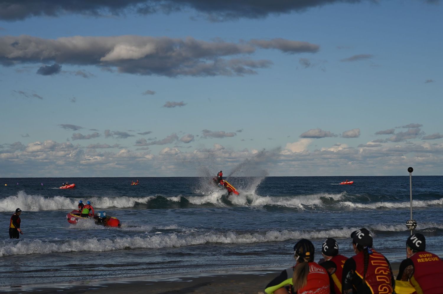 Surf lifesavers compete in ocean waves during a beach rescue competition with colorful boards and red uniforms on a sunny day.