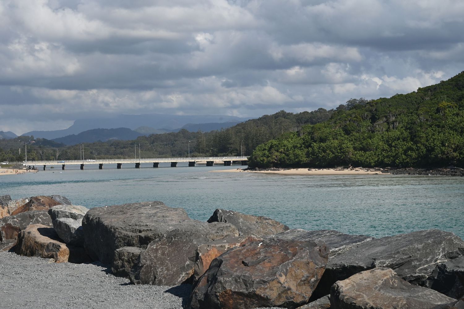 Rocky shoreline with turquoise water, a pier, lush green forest, and mountains under cloudy skies.