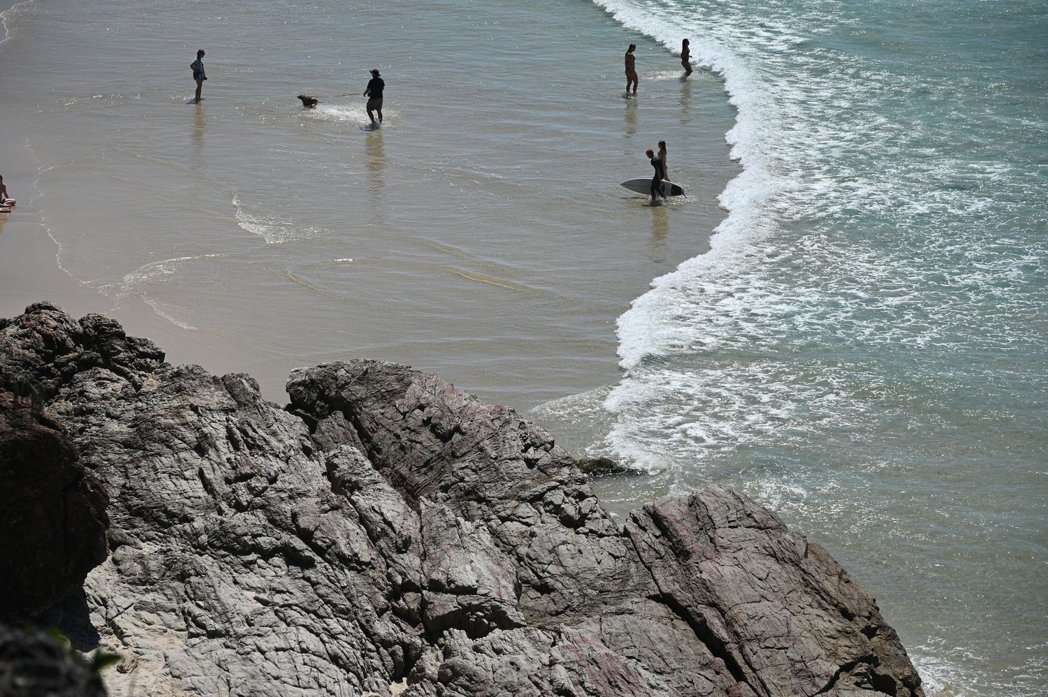 People wading in ocean waves on a rocky beach with clear turquoise water and large stone formations in the foreground.