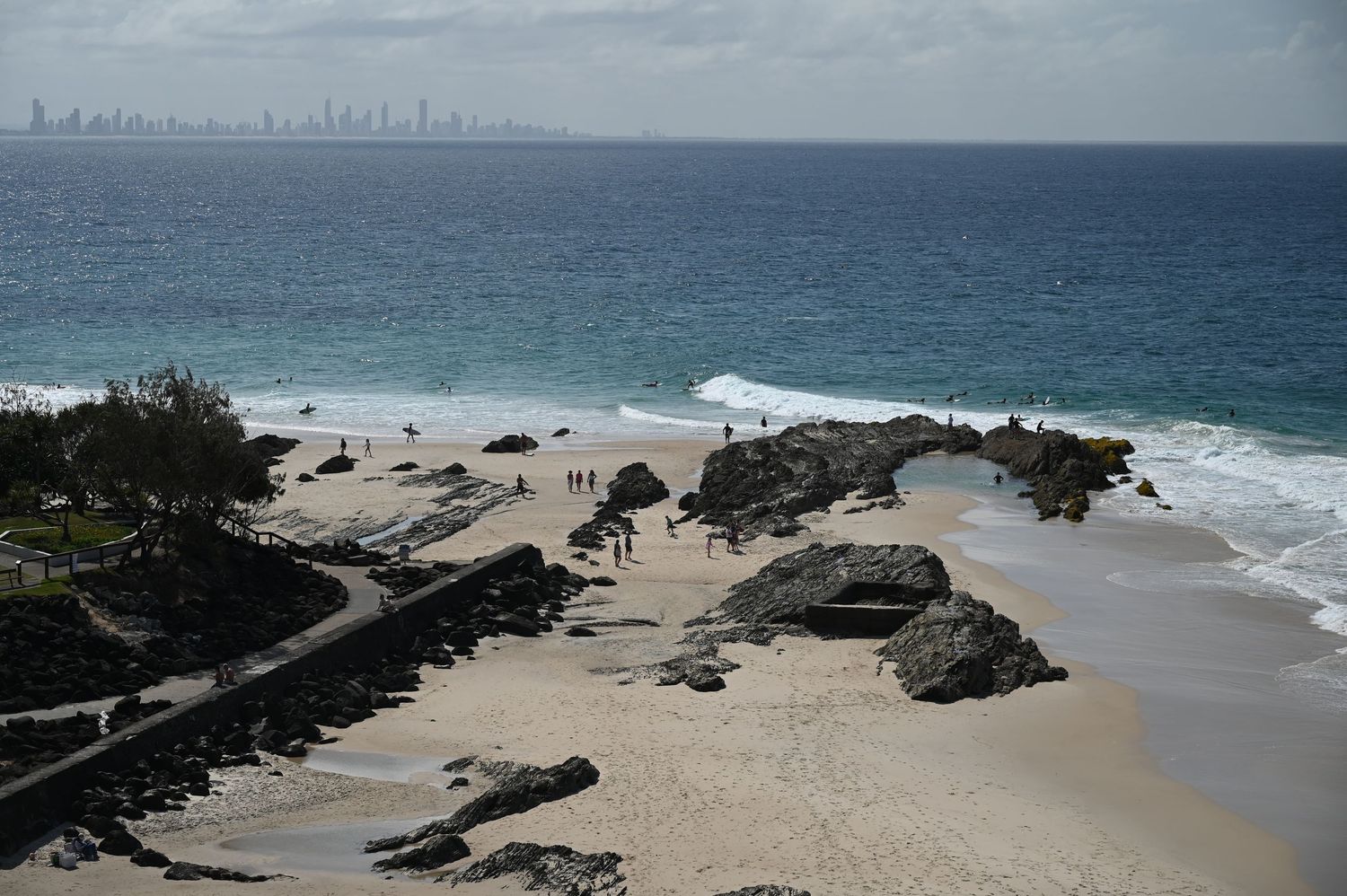 Aerial view of a rocky beach with golden sand, ocean waves, and a city skyline visible in the distance.