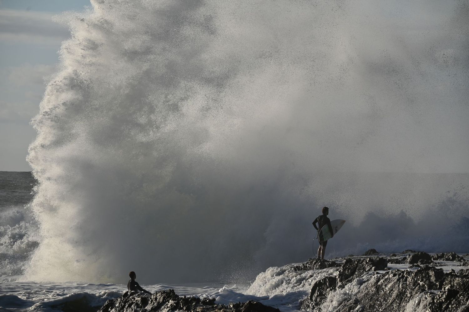 Two surfers stand on rocky shore as massive wave crashes behind them in dramatic ocean scene.