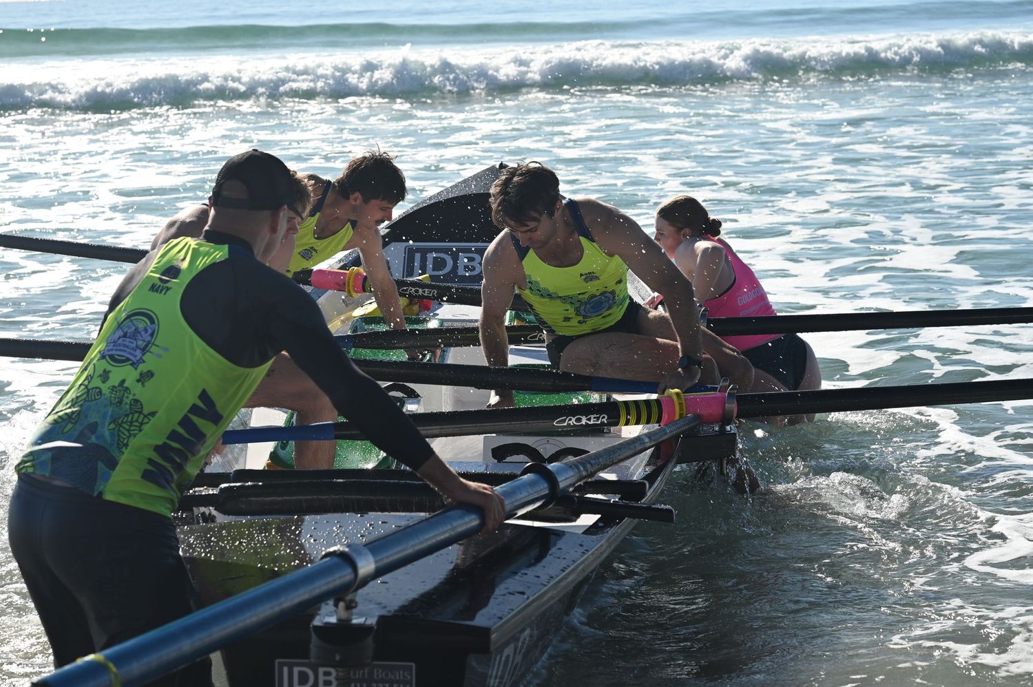 Surf rowing team members in green uniforms maneuvering their boat through ocean waves during a beach competition.
