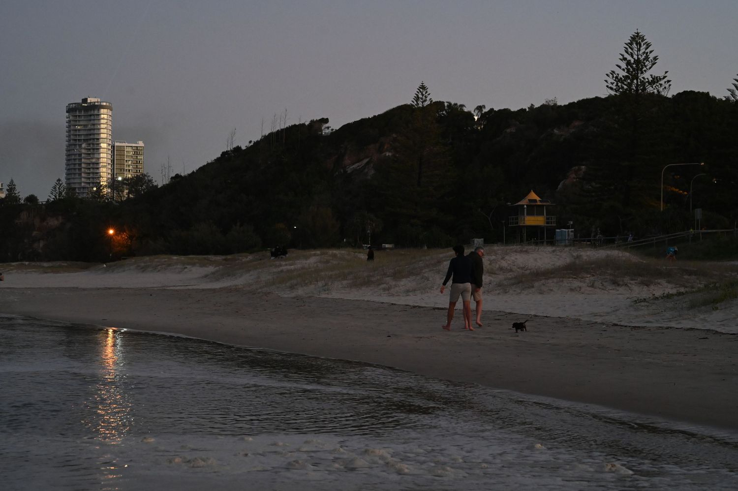 Person walking on a twilight beach with calm waves, tall buildings and lush trees visible in the background.