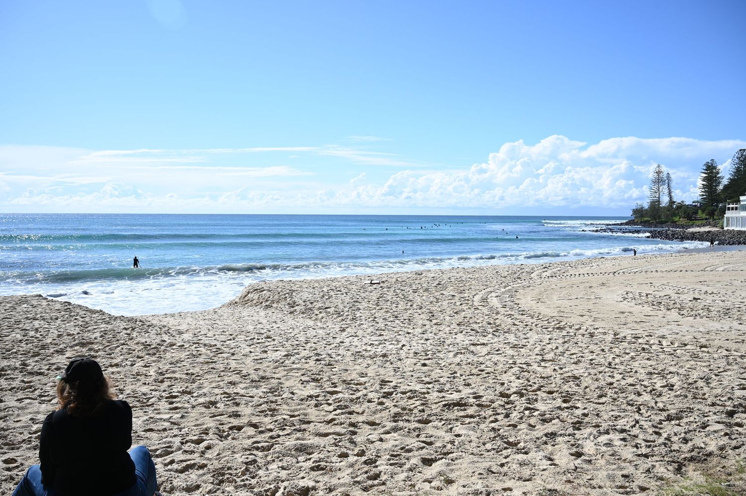 Person sitting on a sandy beach with calm ocean waves and a lighthouse in the background on a sunny day.