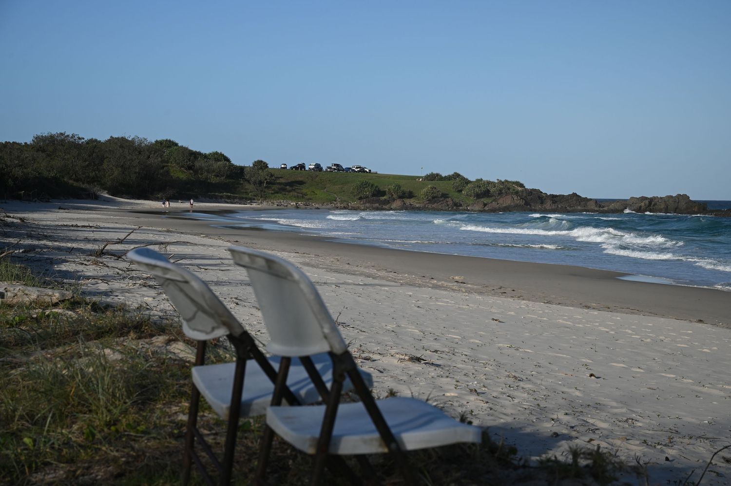 A white folding chair sits on a sandy beach with gentle waves and green headland under a clear blue sky.
