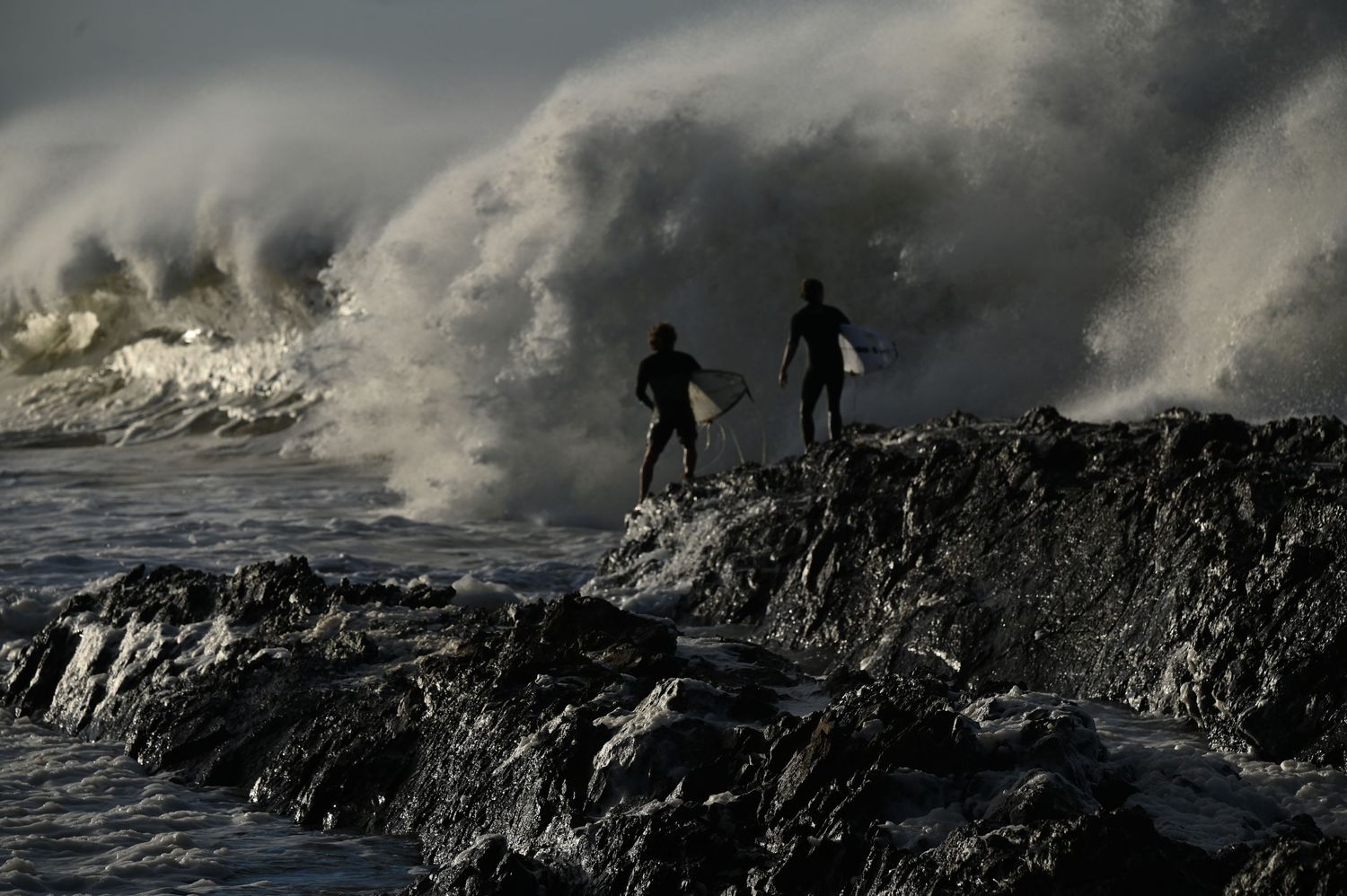 Two surfers stand on rocky shore silhouetted against massive crashing waves in dramatic ocean surf scene.