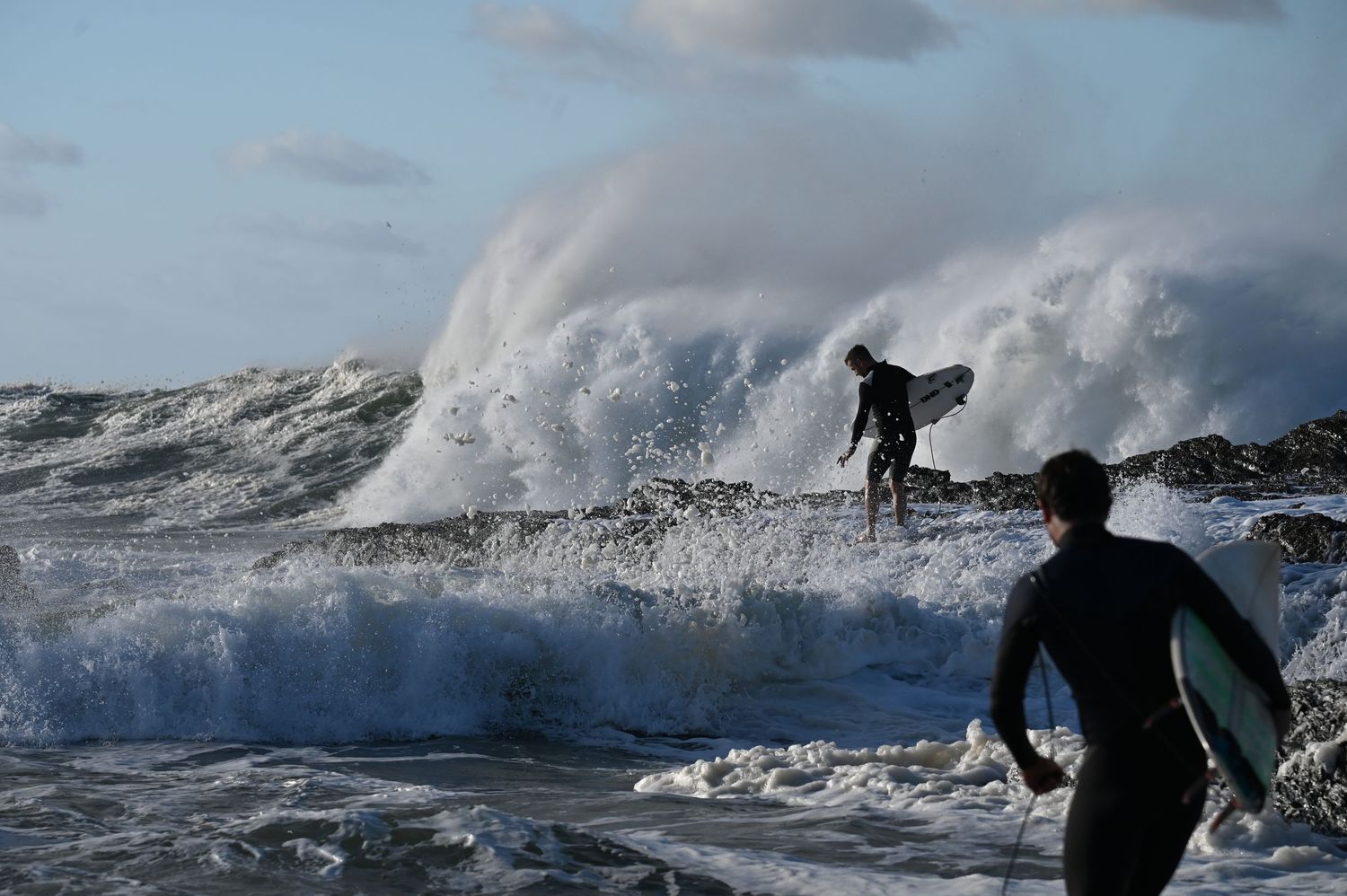 Two surfers in wetsuits brave massive crashing waves on a rocky shoreline under dramatic cloudy skies.
