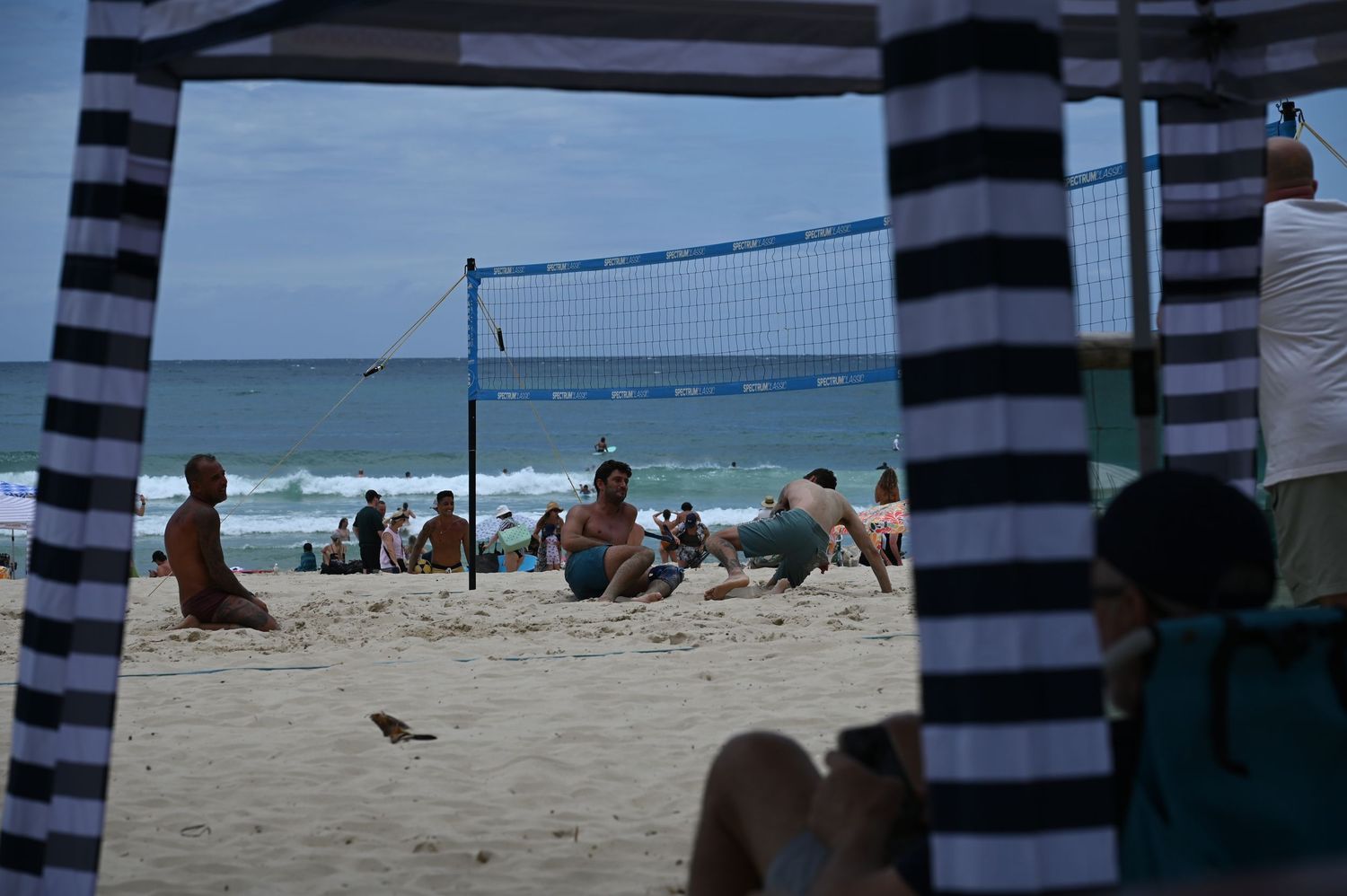 People relaxing on a sandy beach with a volleyball net, ocean waves, and striped canopy framing the scene.