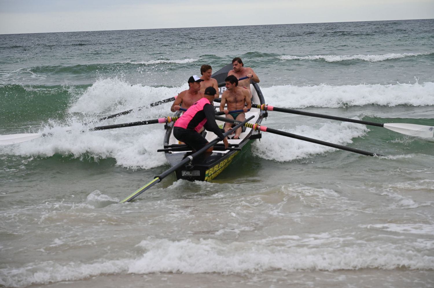 Team of rowers navigating a surf boat through crashing ocean waves during a beach rowing race.