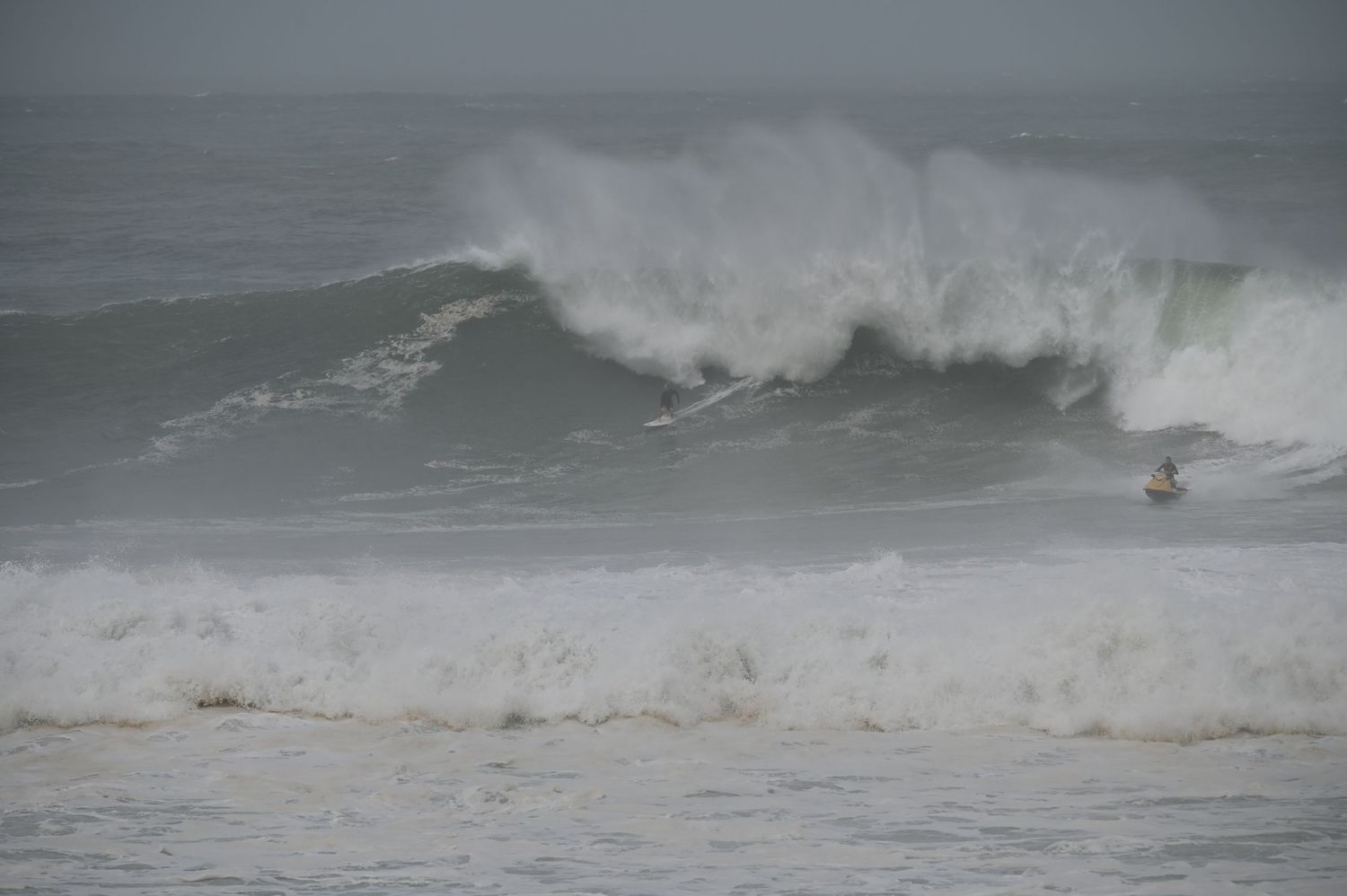 Surfers navigating enormous crashing waves during a stormy grey day at the beach.