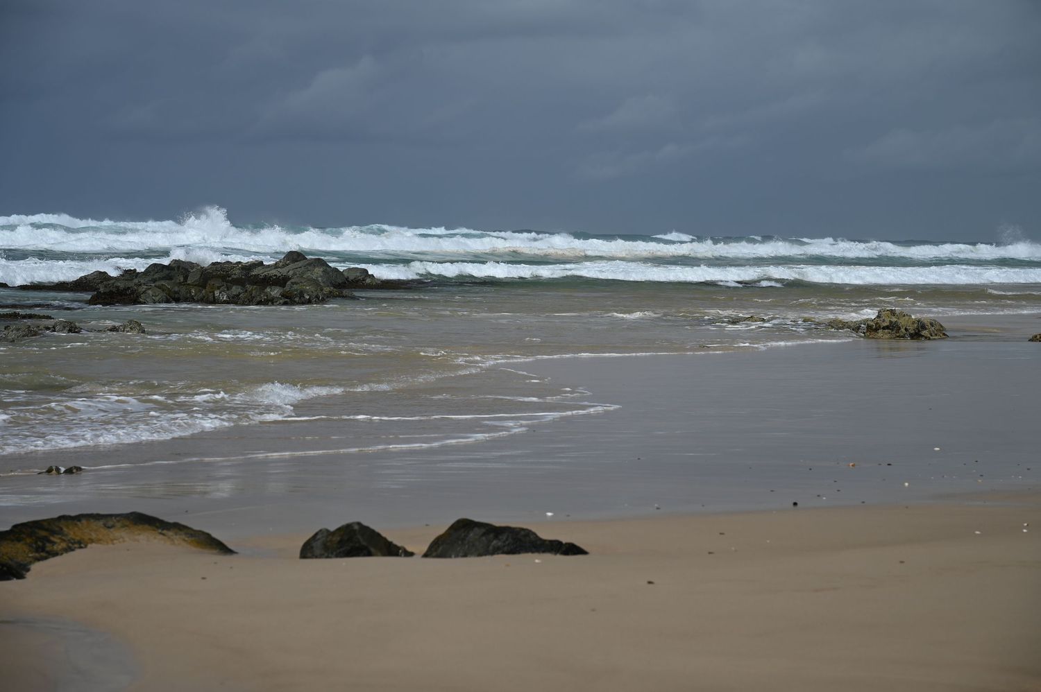 Stormy beach scene with rough ocean waves, dark clouds, sandy shore, and scattered rocks on a dramatic coastline.