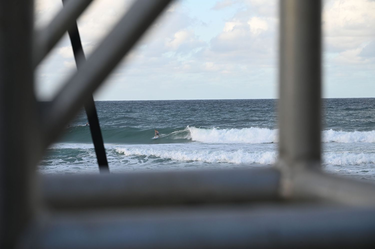 Ocean waves rolling toward shore viewed through white railing frame on a partly cloudy day.