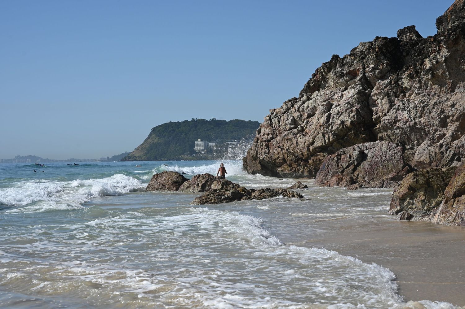 Rocky coastal beach with crashing waves, rugged cliffs, and a scenic headland under a clear blue sky.
