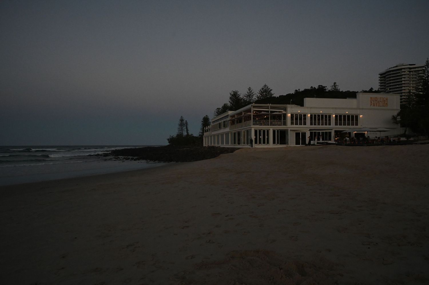 Twilight beachfront scene with a white building restaurant on sandy shore at dusk, calm ocean waves in background.