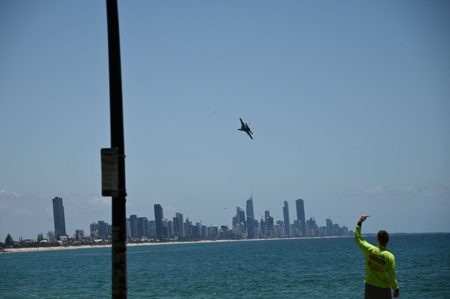 Person in yellow shirt waves at a jet flying over Gold Coast skyline with turquoise ocean in foreground.